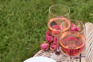 Photo of Picnic basket with glasses of delicious rose wine and flowers outdoors, space for text