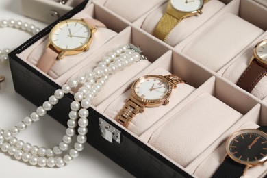 Photo of Jewelry box with many stylish wristwatches and pearl necklace on white table, closeup