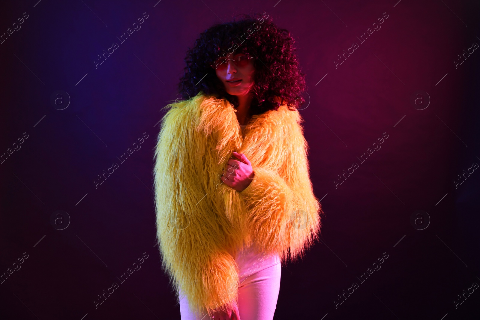 Photo of Beautiful young woman in yellow fur coat and sunglasses on color background in neon lights