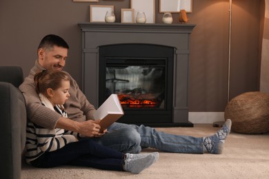 Photo of Happy father and daughter reading book together on floor near fireplace at home