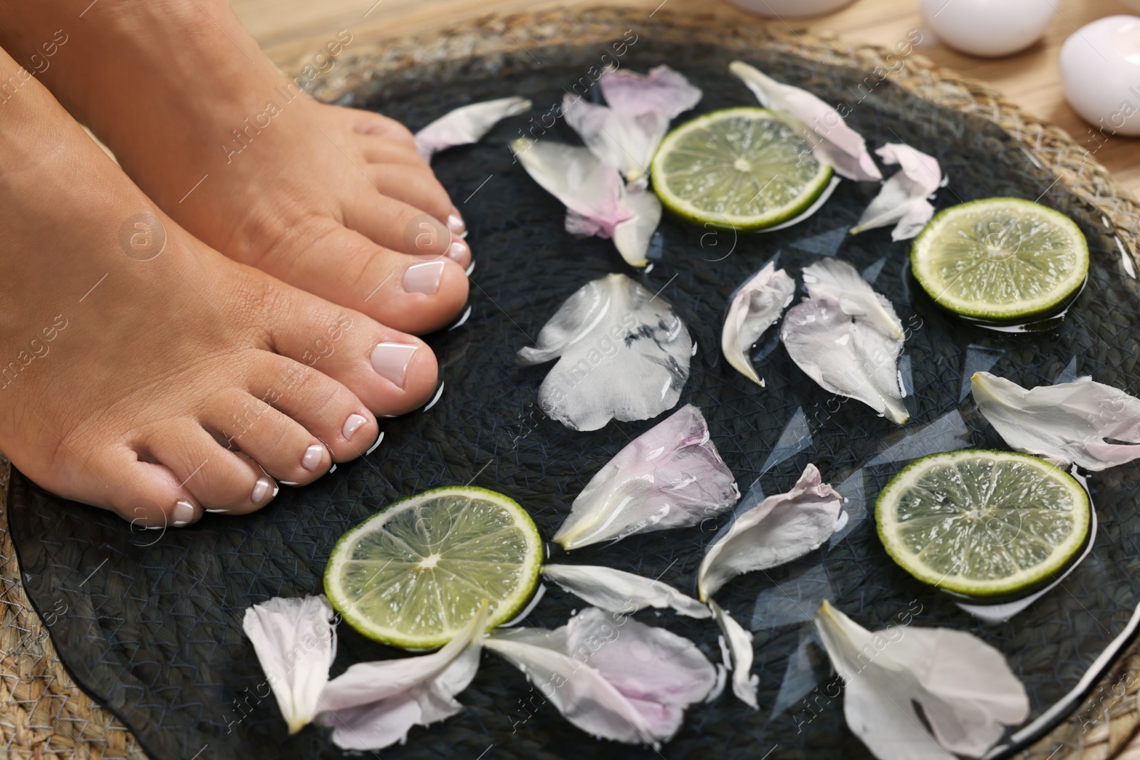 Photo of Woman soaking her feet in plate with water, flower petals and lime slices, closeup. Pedicure procedure