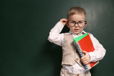 Photo of Cute little child wearing glasses near chalkboard, space for text. First time at school