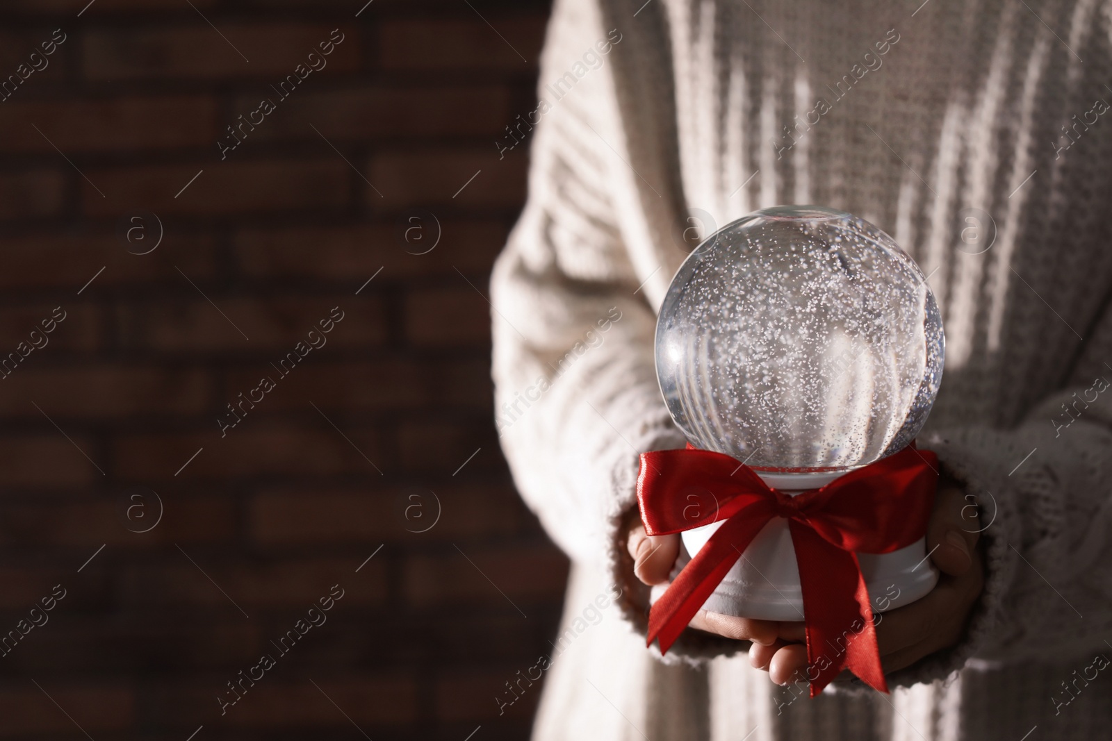 Photo of Woman holding Christmas snow globe with red bow on brick wall background, closeup. Space for text