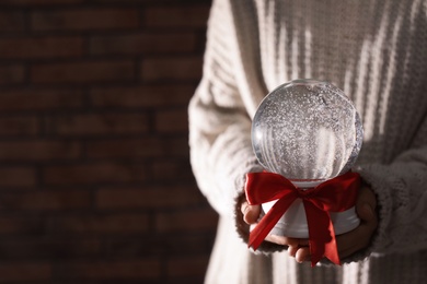 Photo of Woman holding Christmas snow globe with red bow on brick wall background, closeup. Space for text