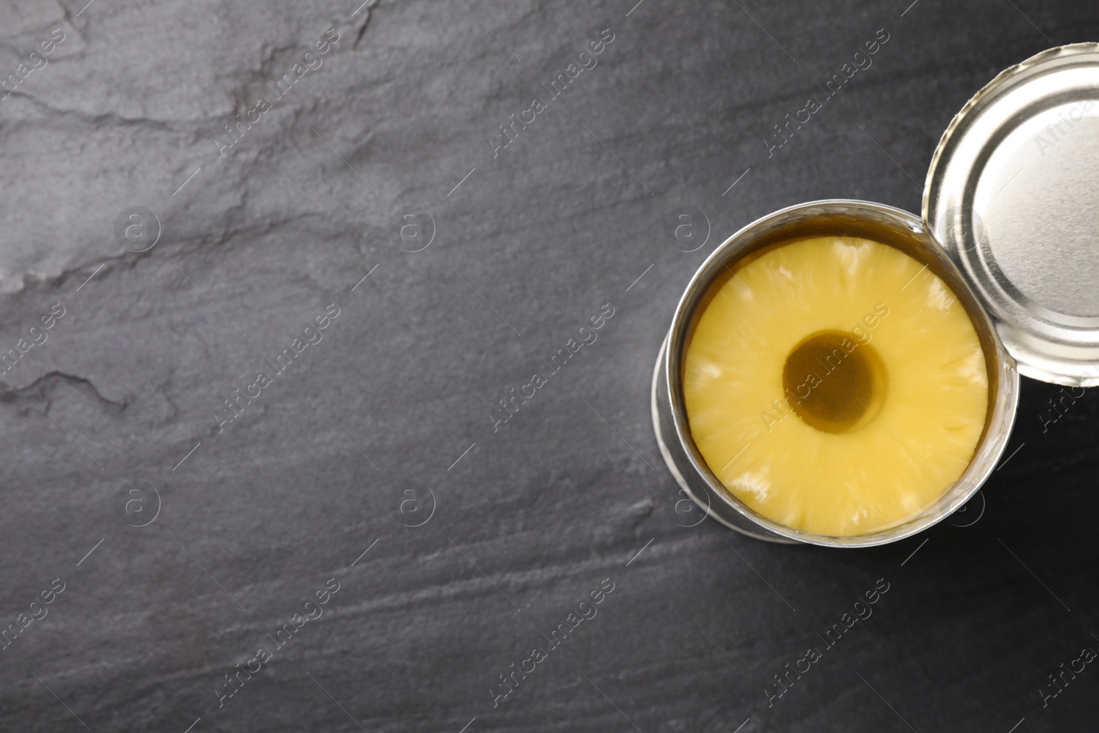 Photo of Tin with canned pineapple on black slate table, top view. Space for text