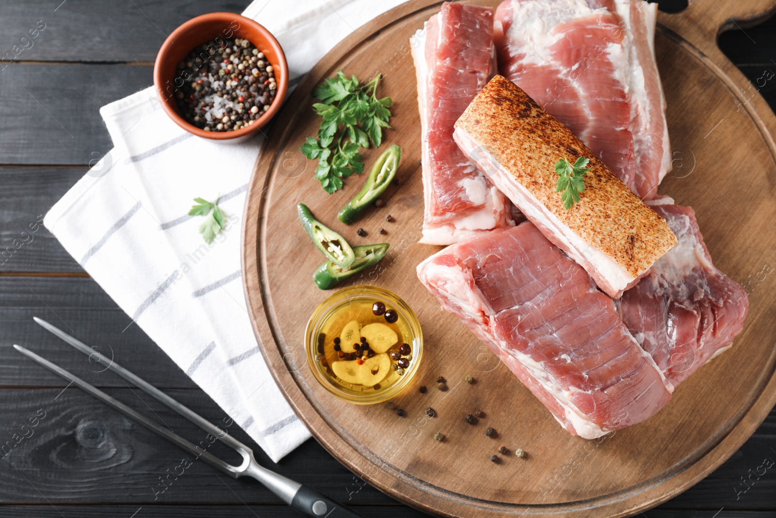 Photo of Pieces of raw pork belly, chili pepper, peppercorns, oil and parsley on black wooden table, top view