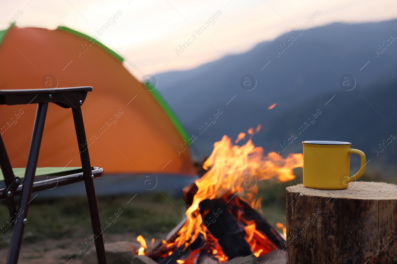 Photo of Yellow mug with hot drink on wooden stump near bonfire outdoors, space for text. Camping season
