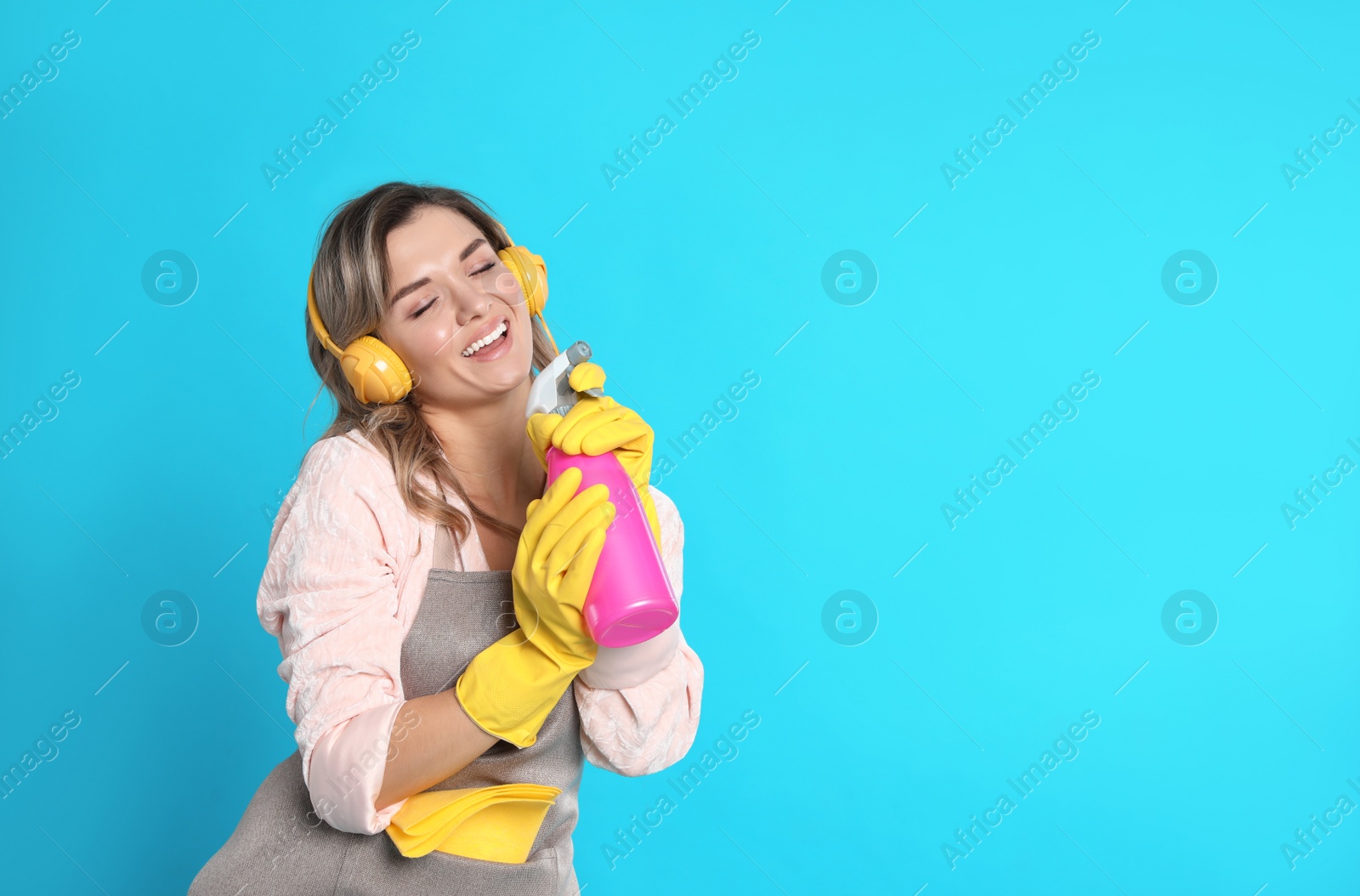 Photo of Beautiful young woman with headphones and bottle of detergent singing on light blue background. Space for text