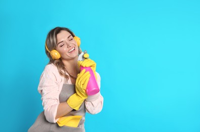Beautiful young woman with headphones and bottle of detergent singing on light blue background. Space for text