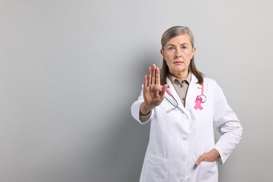 Photo of Mammologist with pink ribbon showing stop gesture on gray background, space for text. Breast cancer awareness