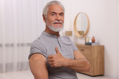Photo of Senior man with adhesive bandage on his arm after vaccination showing thumb up at home