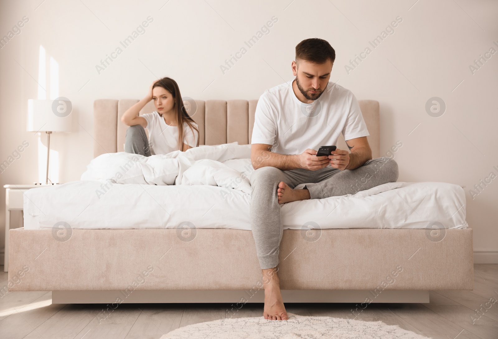 Photo of Young man preferring smartphone over his girlfriend on bed at home. Relationship problems
