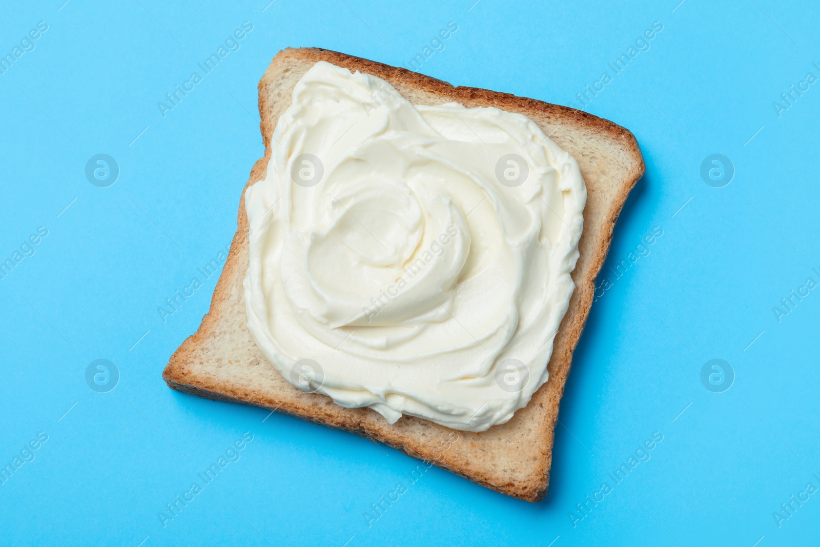 Photo of Slice of bread with tasty cream cheese on light blue background, top view