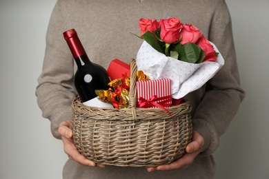 Photo of Man holding wicker basket with gifts, bouquet and wine on grey background, closeup