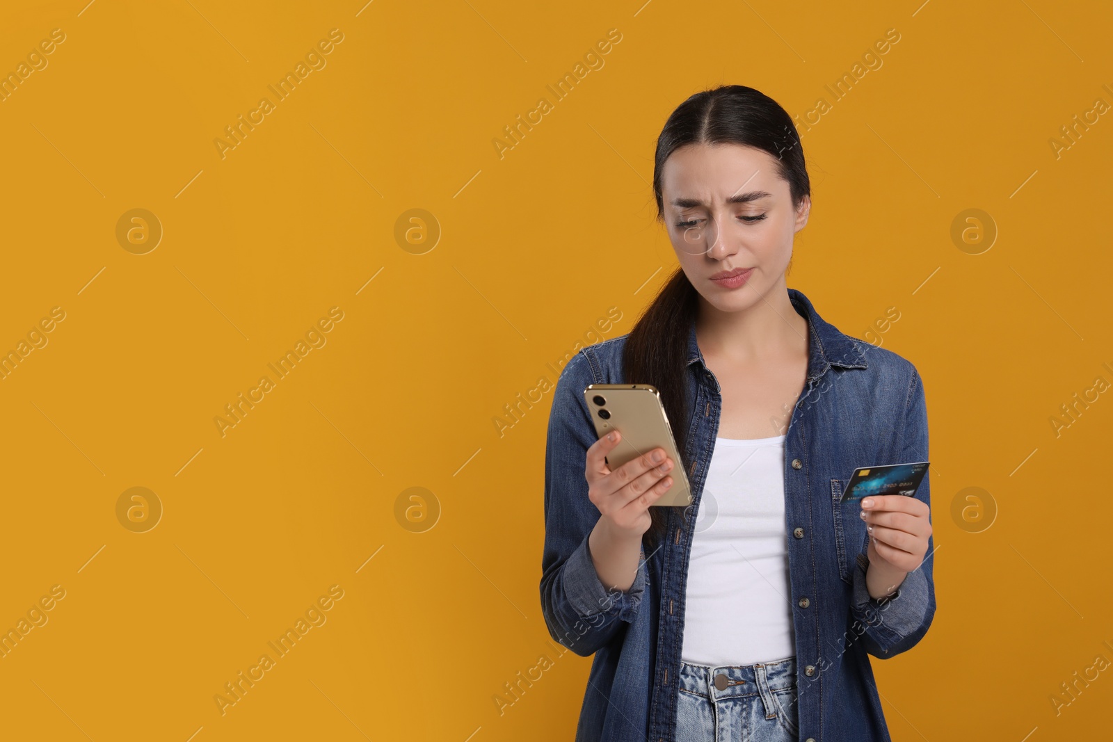 Photo of Confused woman with credit card and smartphone on orange background, space for text. Debt problem