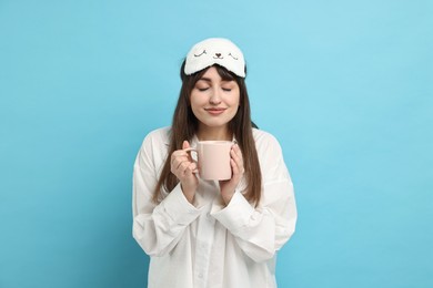 Photo of Woman in pyjama and sleep mask holding cup of drink on light blue background