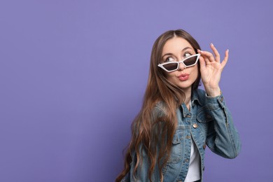 Photo of Beautiful young woman with sunglasses blowing kiss on purple background, space for text
