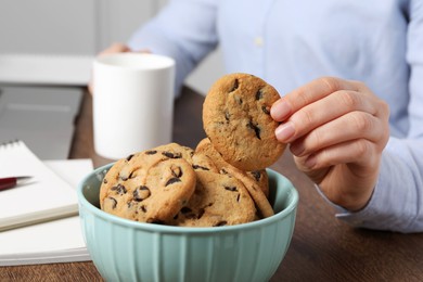 Photo of Office worker with cup of drink taking chocolate chip cookie from bowl at workplace, closeup