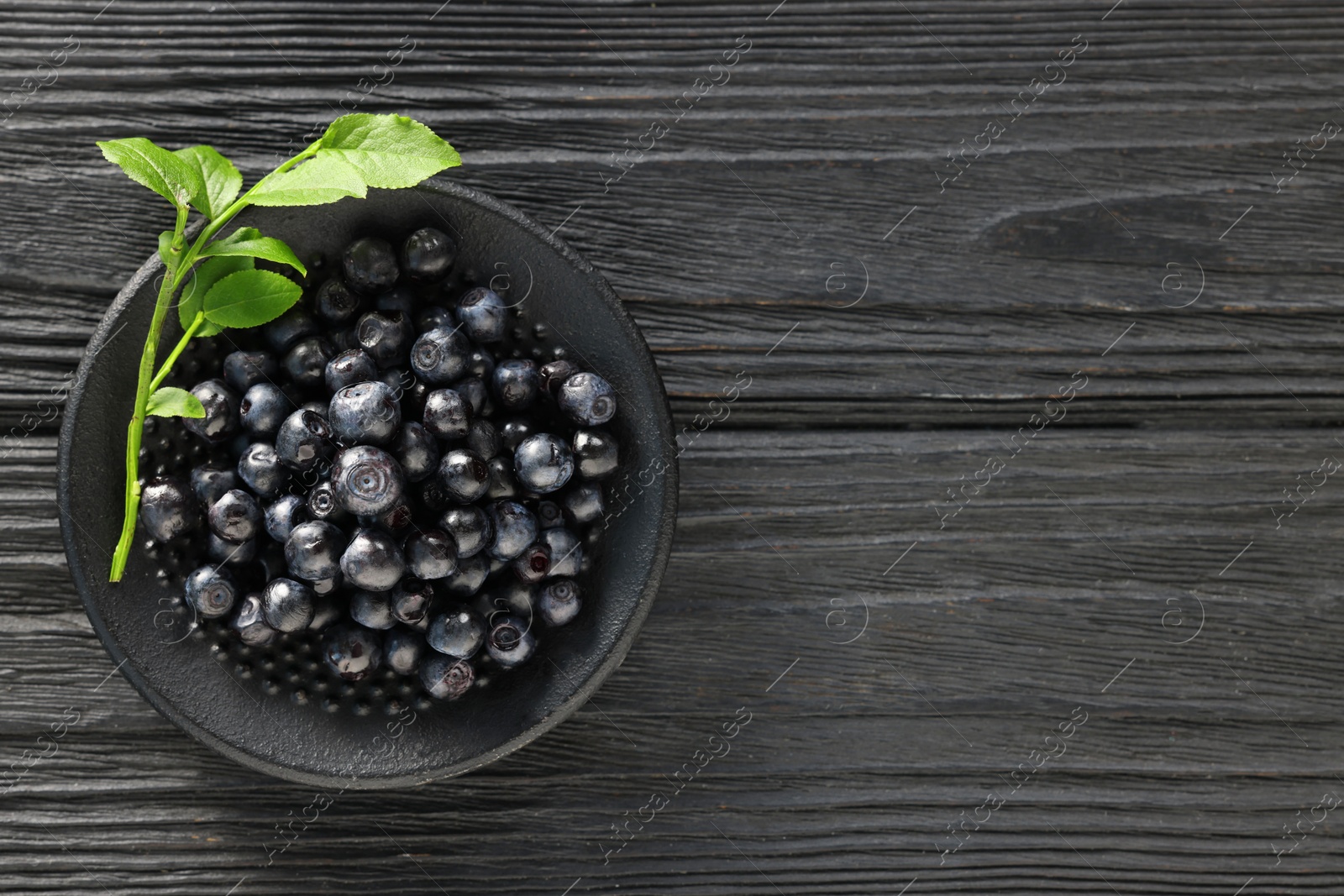 Photo of Tasty fresh bilberries with green leaves in bowl on black wooden table, top view. Space for text