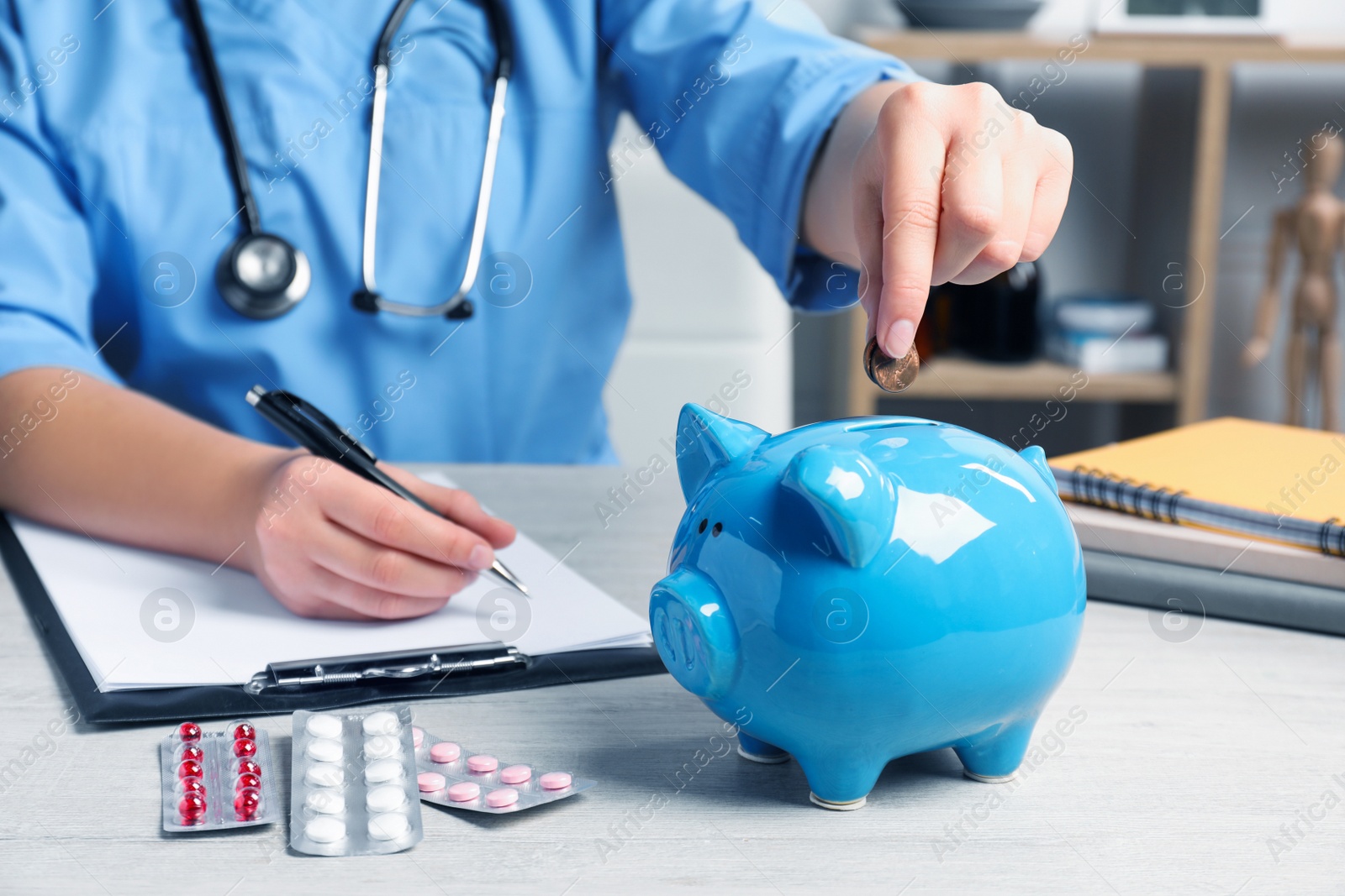 Photo of Doctor putting coins into piggy bank near pills at wooden table in hospital, closeup. Medical insurance