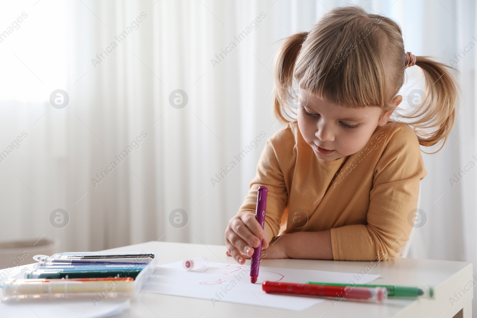 Photo of Cute little girl drawing with marker at white table indoors. Child`s art