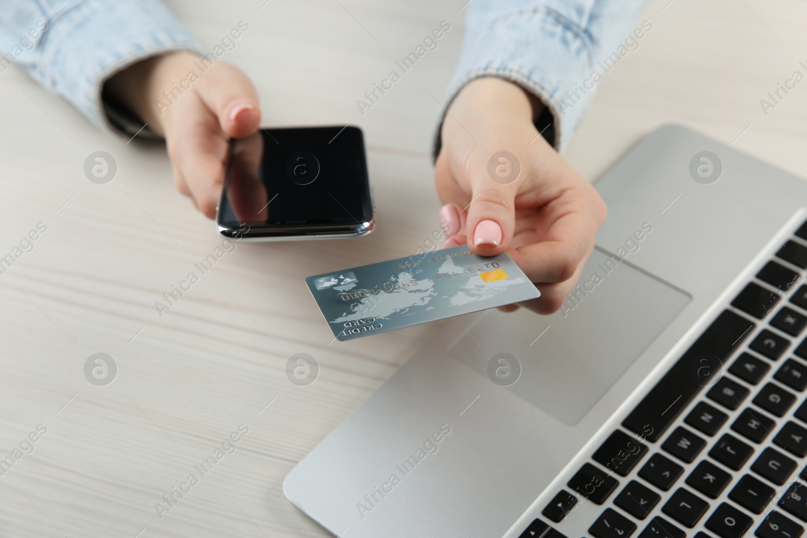 Photo of Online payment. Woman using credit card and smartphone near laptop at white wooden table, closeup