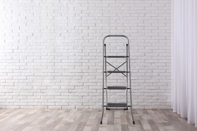 Photo of Modern metal stepladder near white brick wall indoors. Space for text