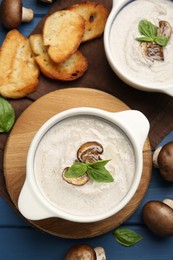 Photo of Delicious mushroom soup in ceramic pots and fresh ingredients on blue wooden table, flat lay