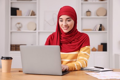 Muslim woman in hijab using laptop at wooden table in room