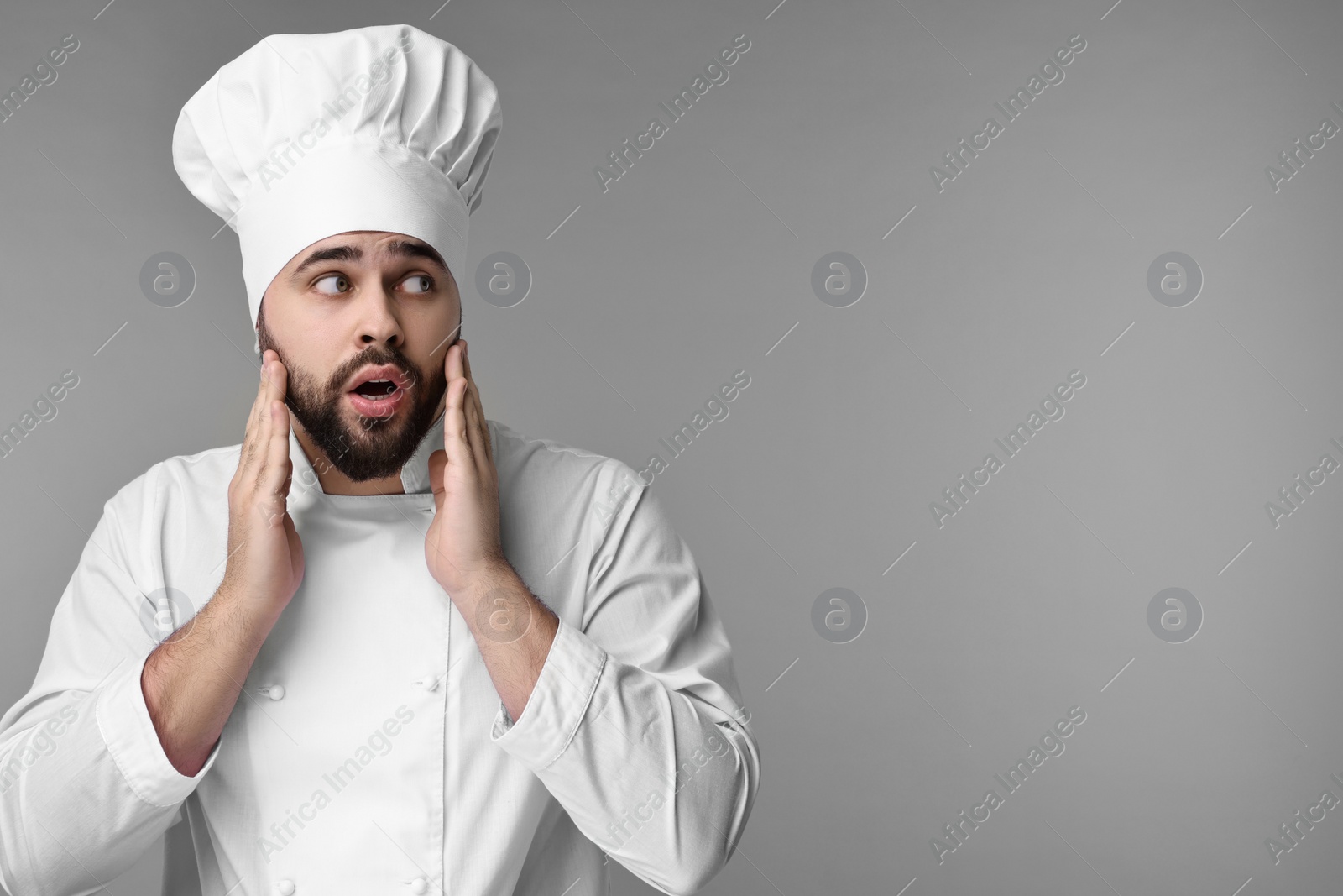 Photo of Surprised young chef in uniform on grey background. Space for text