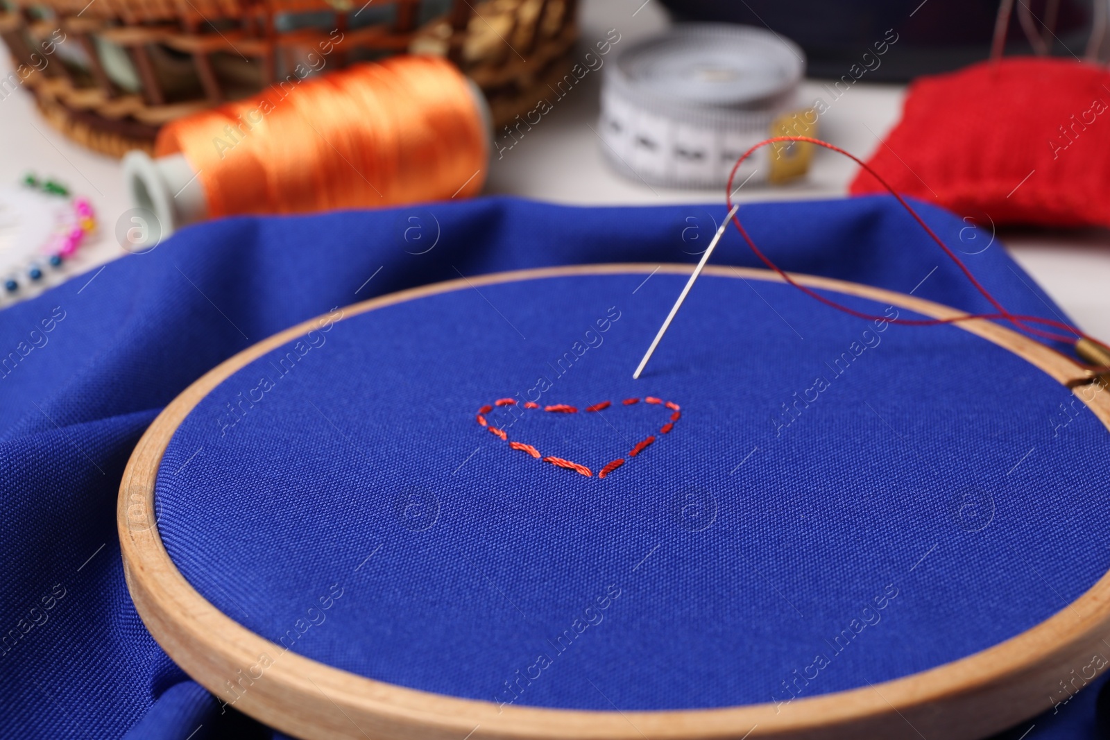 Photo of Red embroidered heart, thread and sewing needle on blue cloth with hoop, closeup