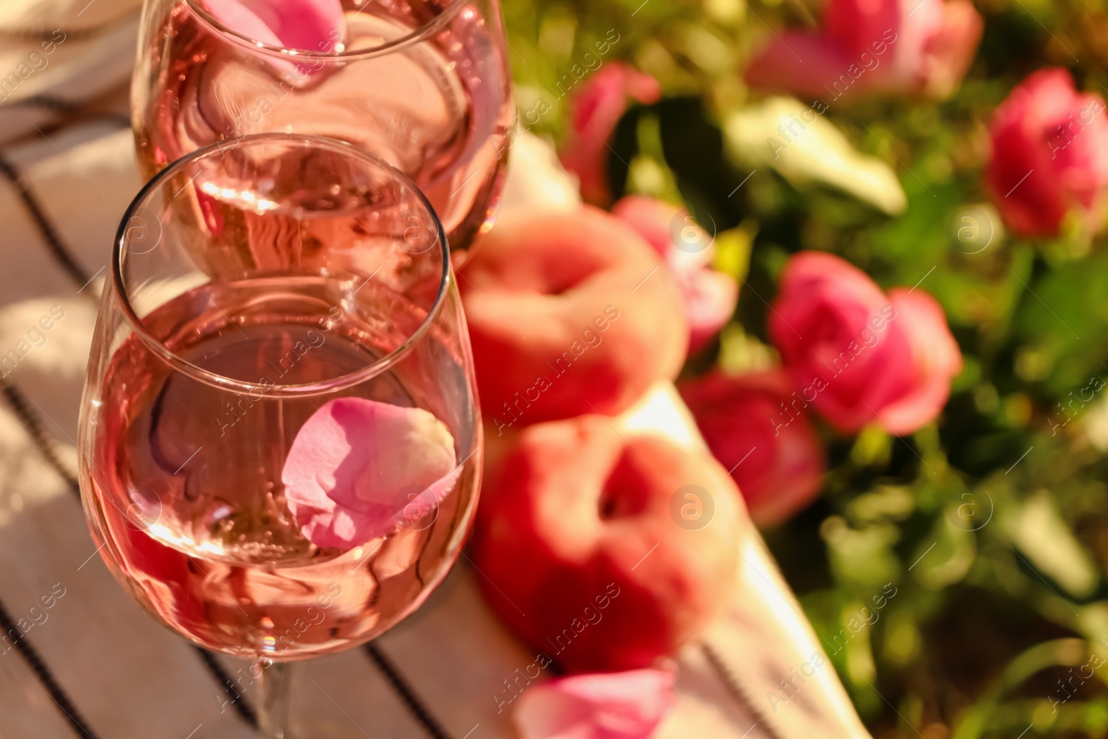 Photo of Glasses of delicious rose wine with petals on white picnic blanket outside. Space for text