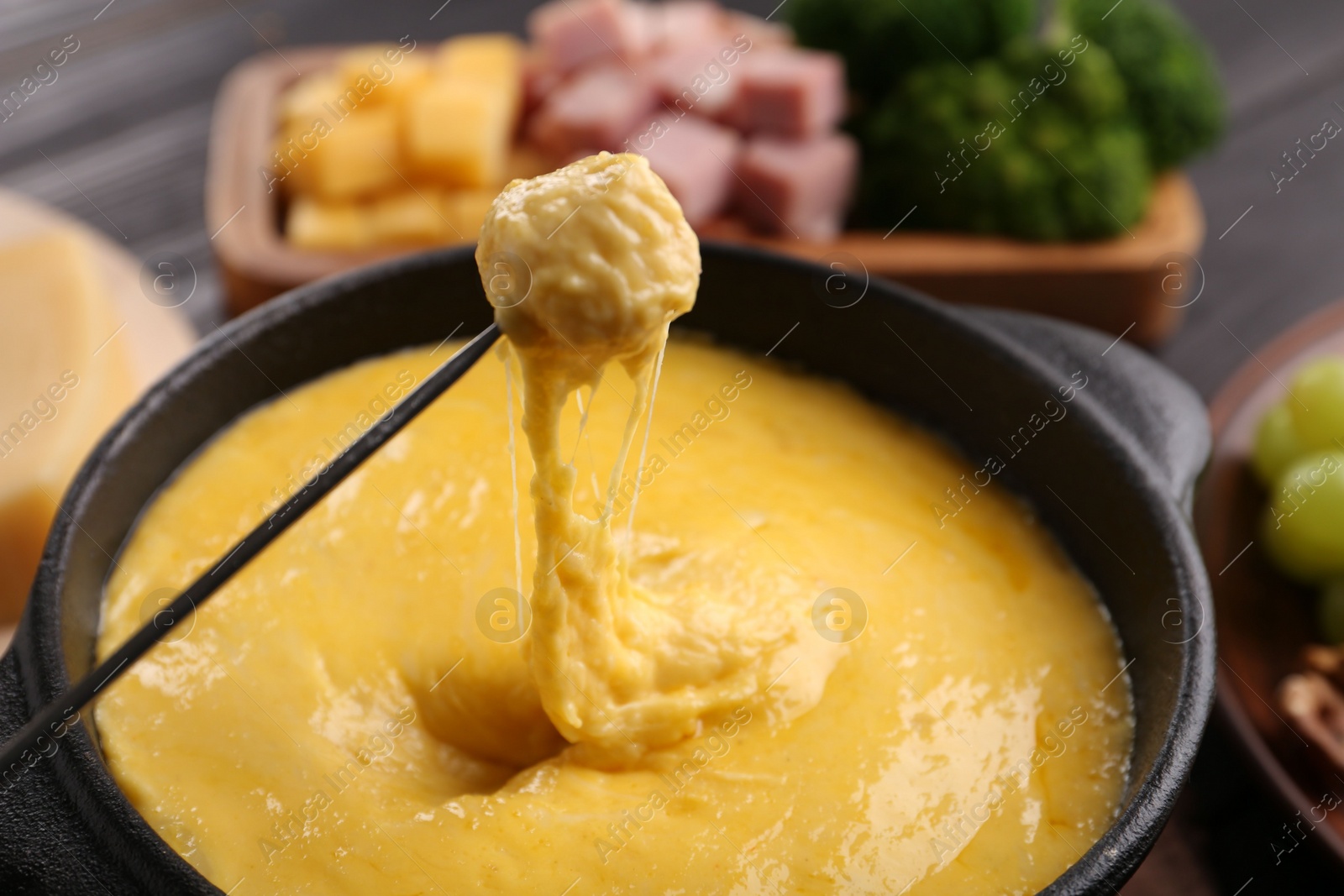 Photo of Dipping piece of ham into fondue pot with melted cheese on table, closeup