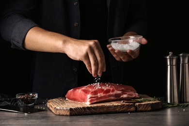 Photo of Man salting fresh raw meat on table against dark background, closeup