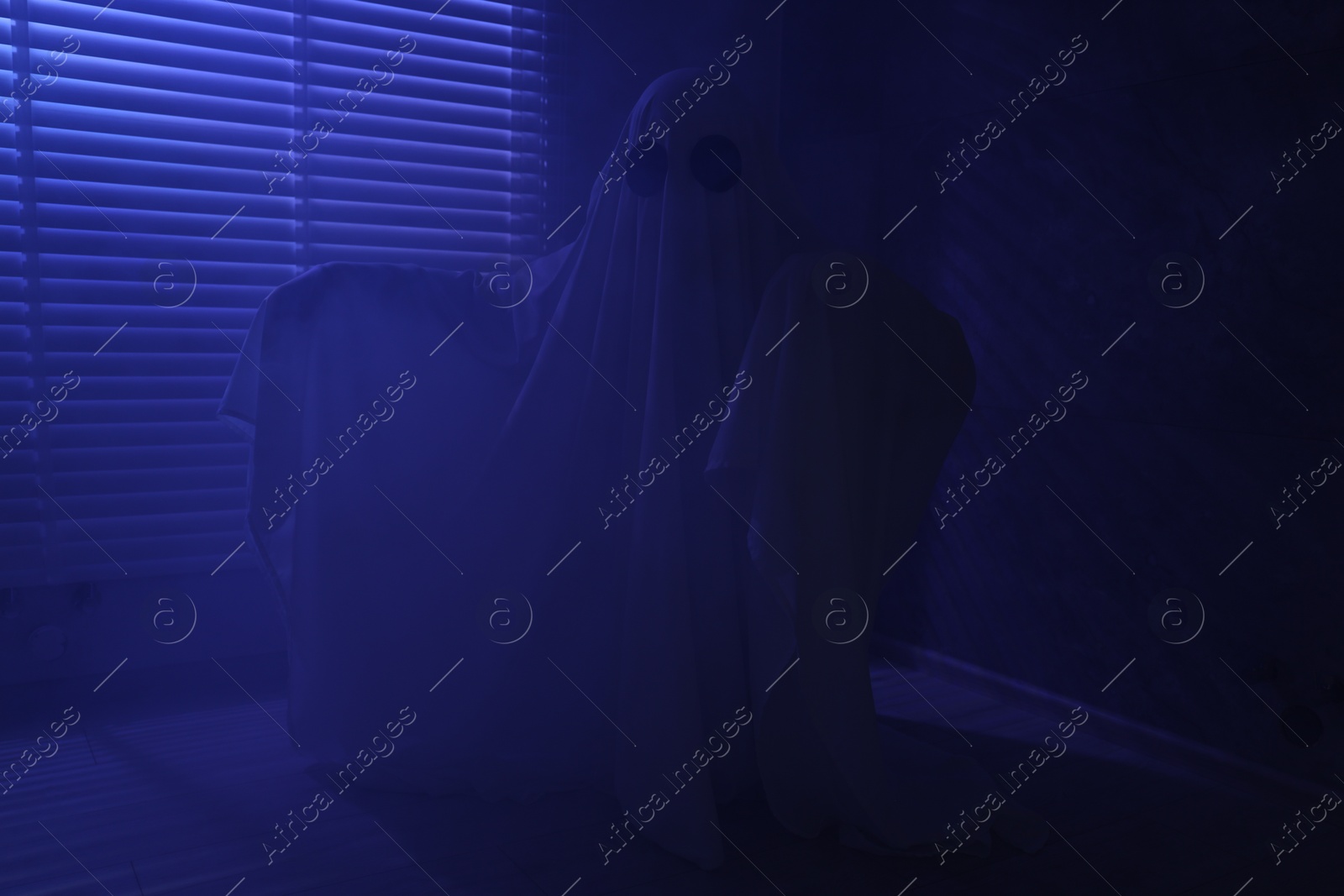 Photo of Creepy ghost. Woman covered with sheet near window in blue light