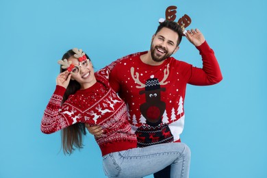 Photo of Happy young couple in Christmas sweaters, reindeer headband and funny glasses on light blue background