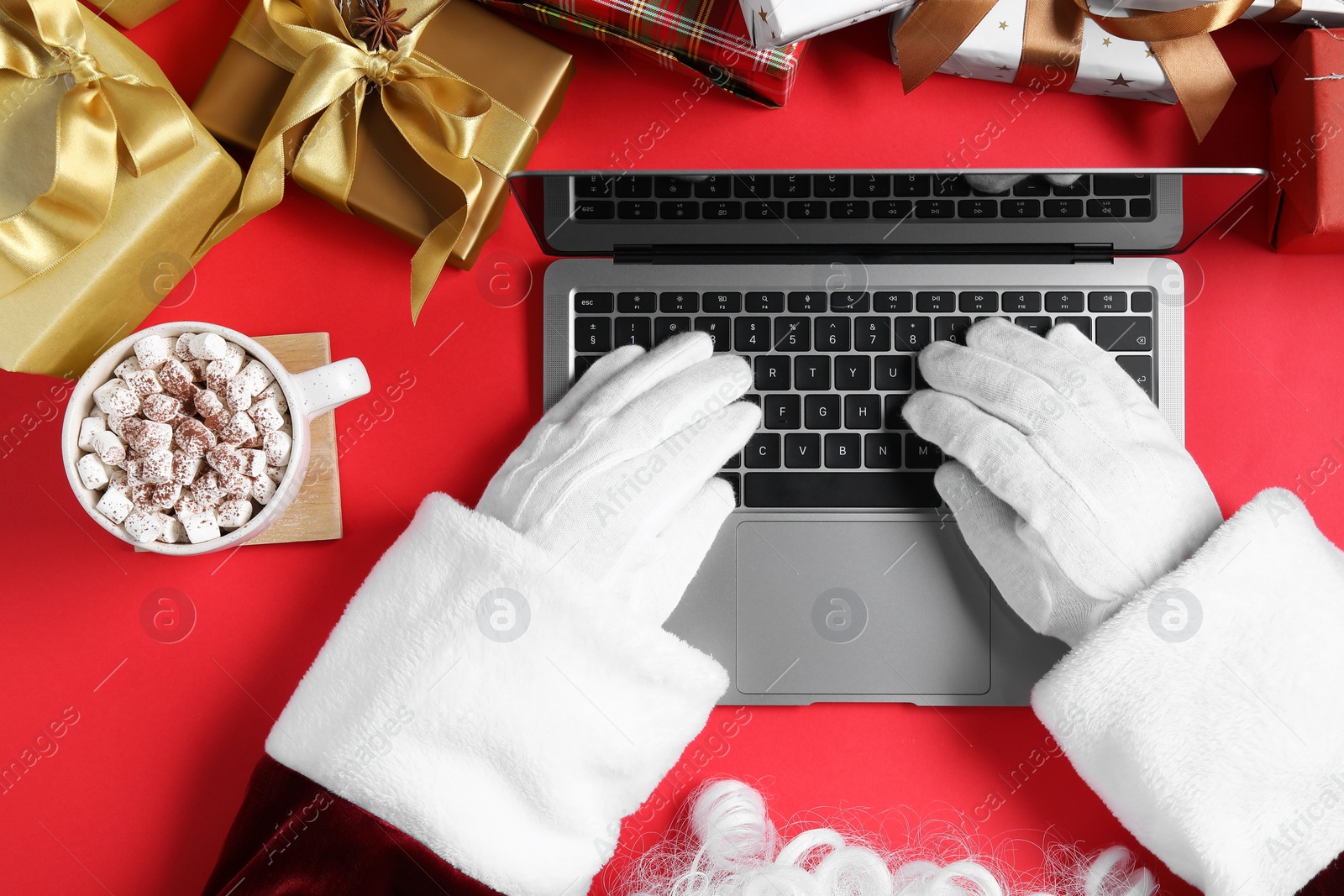 Photo of Santa Claus using laptop, closeup. Gift boxes, cup of drink and Christmas decor on red background, top view