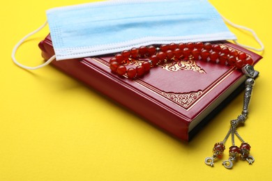 Photo of Muslim prayer beads, Quran and medical mask on yellow background, closeup