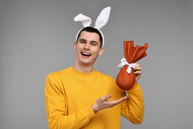 Photo of Easter celebration. Handsome young man with bunny ears holding wrapped gift on grey background