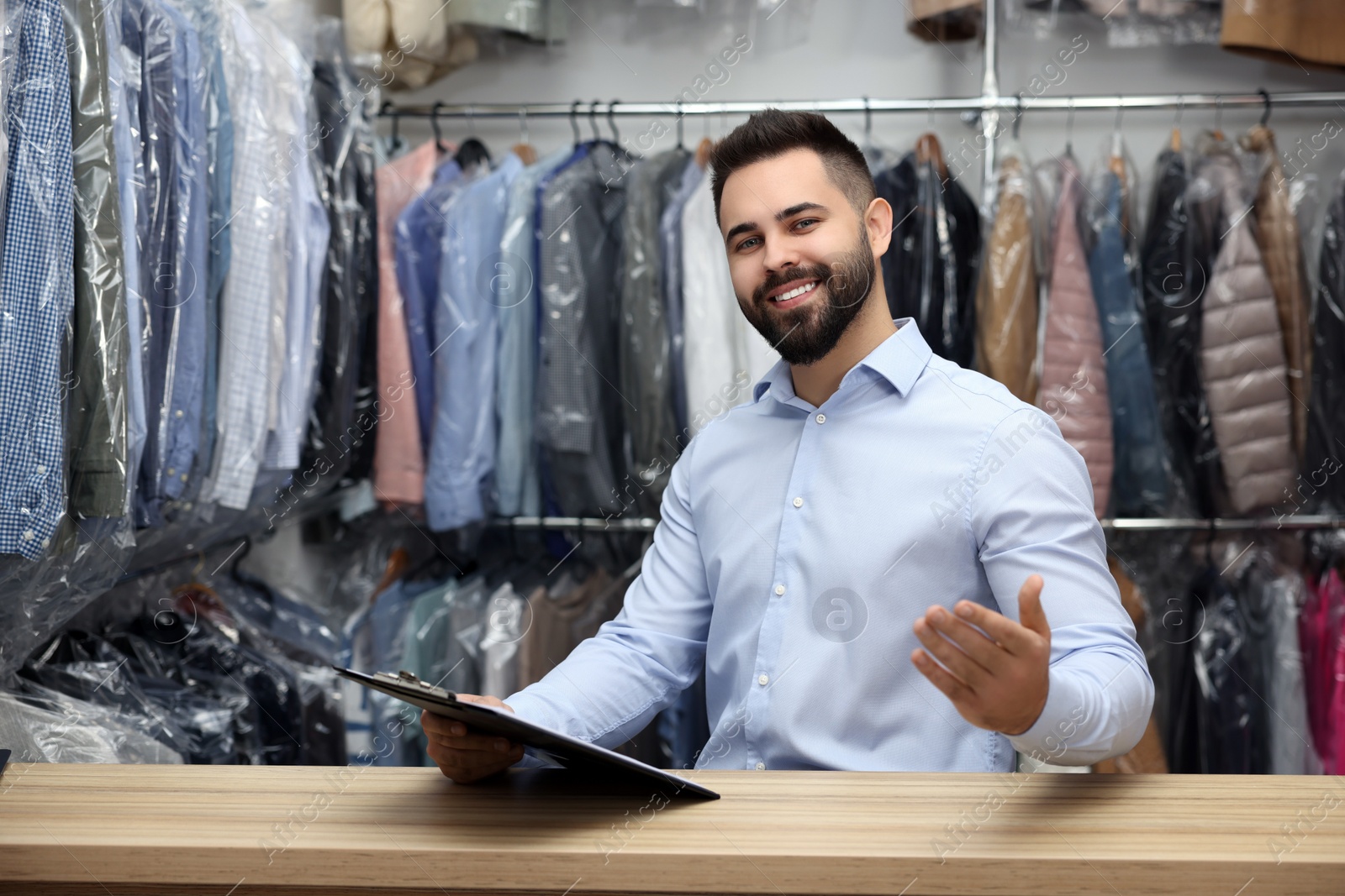 Photo of Dry-cleaning service. Happy worker holding clipboard at counter indoors