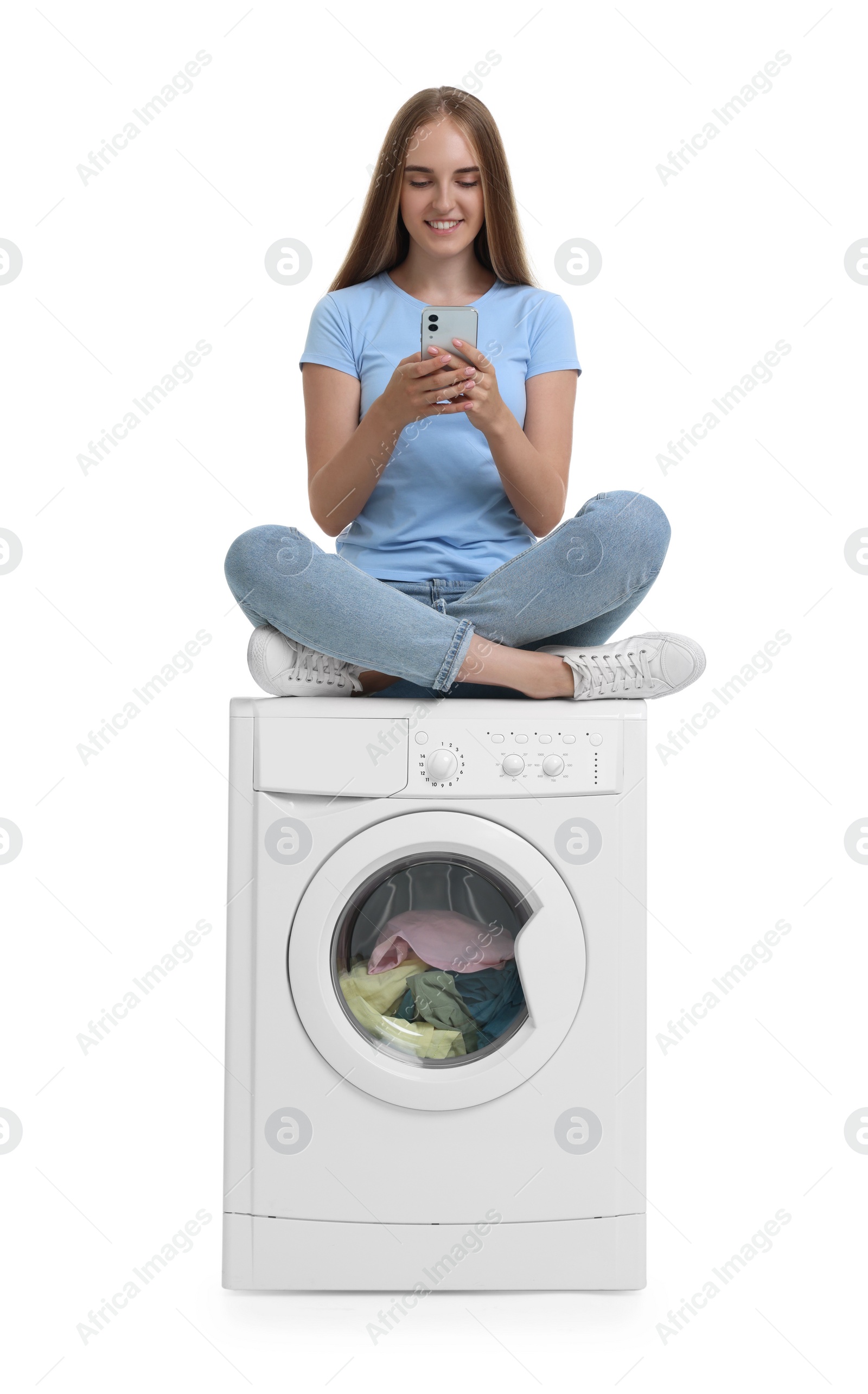 Photo of Beautiful young woman using smartphone on washing machine with laundry against white background