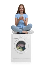 Photo of Beautiful young woman using smartphone on washing machine with laundry against white background