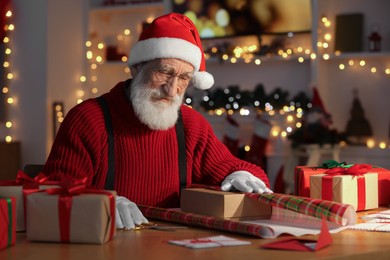 Photo of Santa Claus wrapping gift at his workplace in room decorated for Christmas