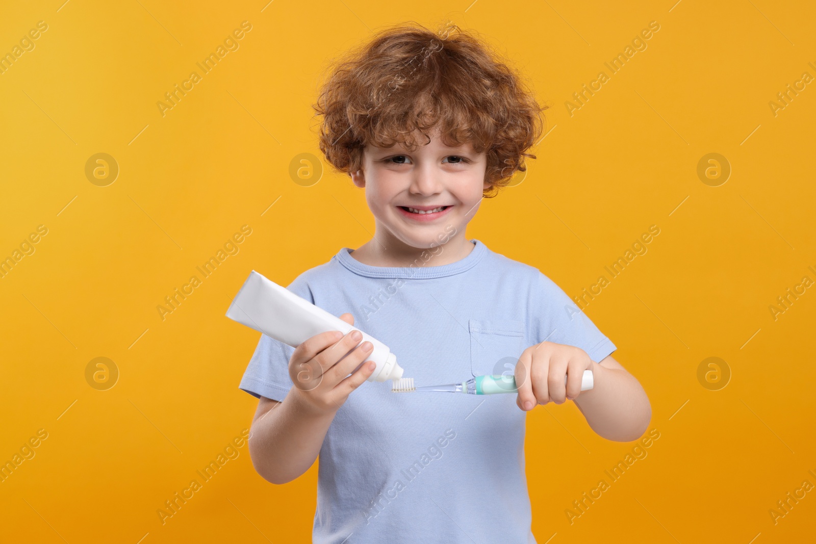 Photo of Cute little boy squeezing toothpaste from tube onto electric toothbrush on yellow background