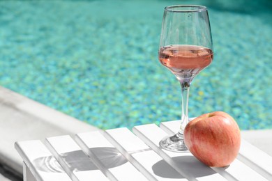 Photo of Glass of tasty rose wine and apple on wooden table near swimming pool, space for text