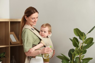 Photo of Mother spraying houseplant with water while holding her child in sling (baby carrier) at home