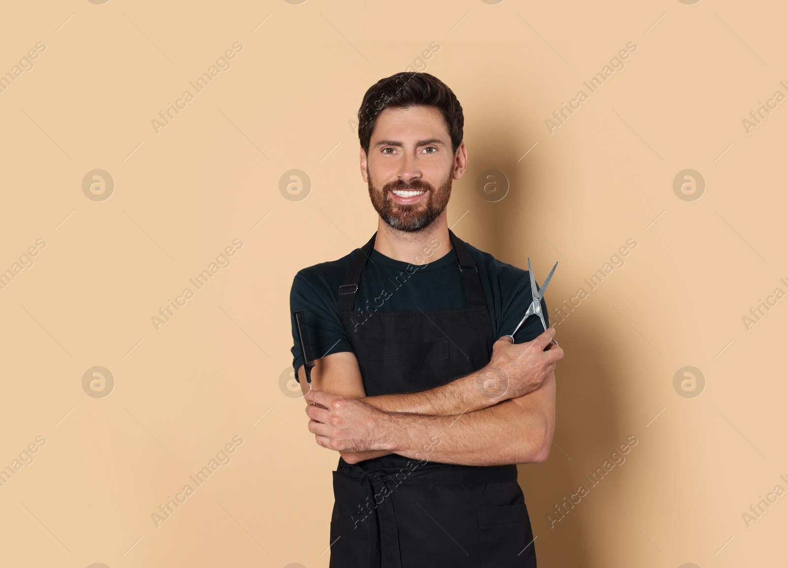 Photo of Smiling hairdresser in apron holding comb and scissors on light brown background