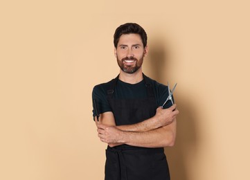 Photo of Smiling hairdresser in apron holding comb and scissors on light brown background