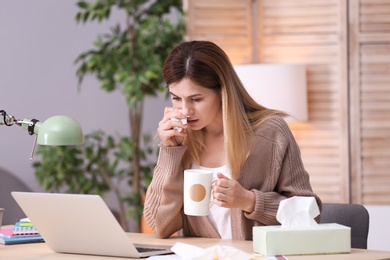 Photo of Sad exhausted woman with tissue and cup of hot drink suffering from cold while working with laptop at table