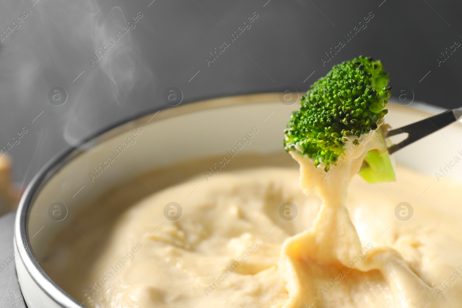 Photo of Dipping piece of broccoli into fondue pot with melted cheese on grey background, closeup